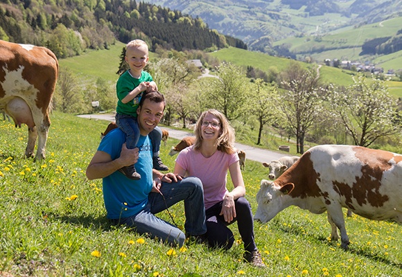 Familie sitzt auf einer Wiese. Im Hintergrund sind Kühe und Berge zu sehen.