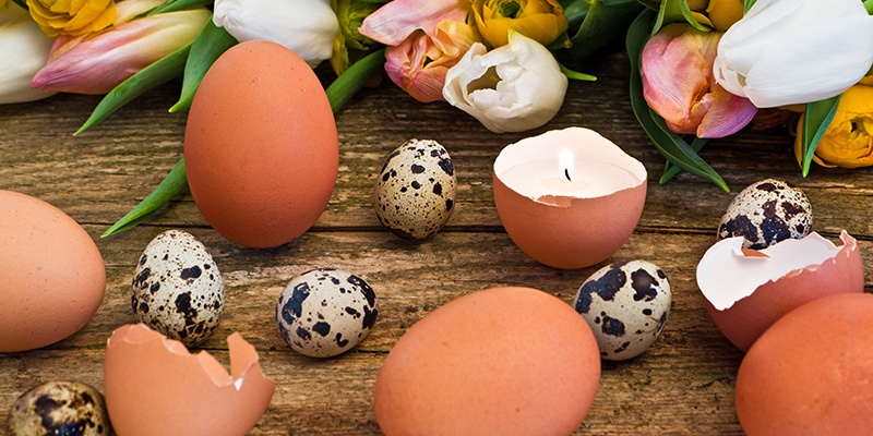 Ganze Wachteleier und Hühnereier auf Holzbrett mit Blumen im Hintergrund und einer Kerze in einer halben Eierschale