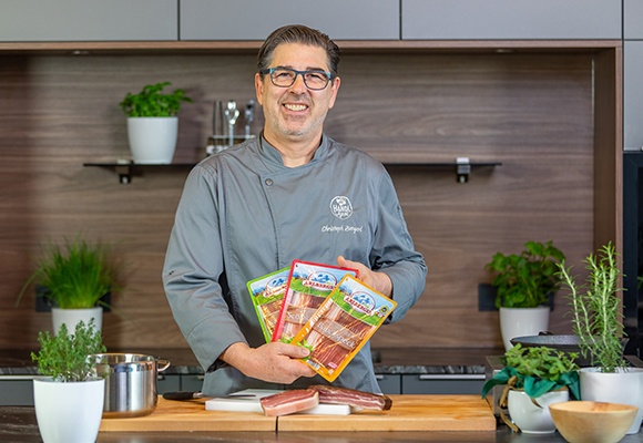 Ein Herr lächelt in die Kamera mit Schinkenspeck in den Händen