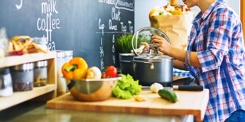 Küchenzeile mit einer Schale Gemüse und einer Frau, die den Deckel eines Kochtopfes anhebt.
