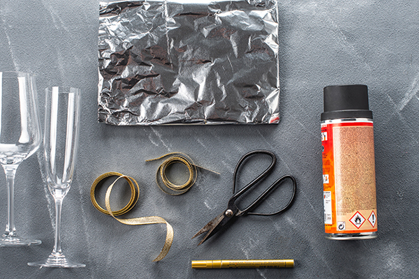 Sektglas, schwarze Sprayflasche, ein goldener Lackstift, Alufolie und eine Schere sind auf einem grauen Hintergrund