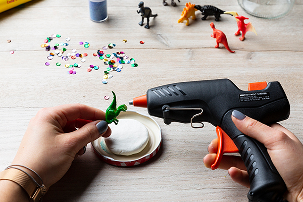 Dinosaurier wird mit Heißklebepistole auf einen Deckel geklebt.
