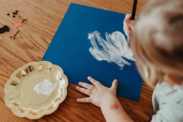 Kleinkind bemalt ein blaues Papier mit weißer Farbe.