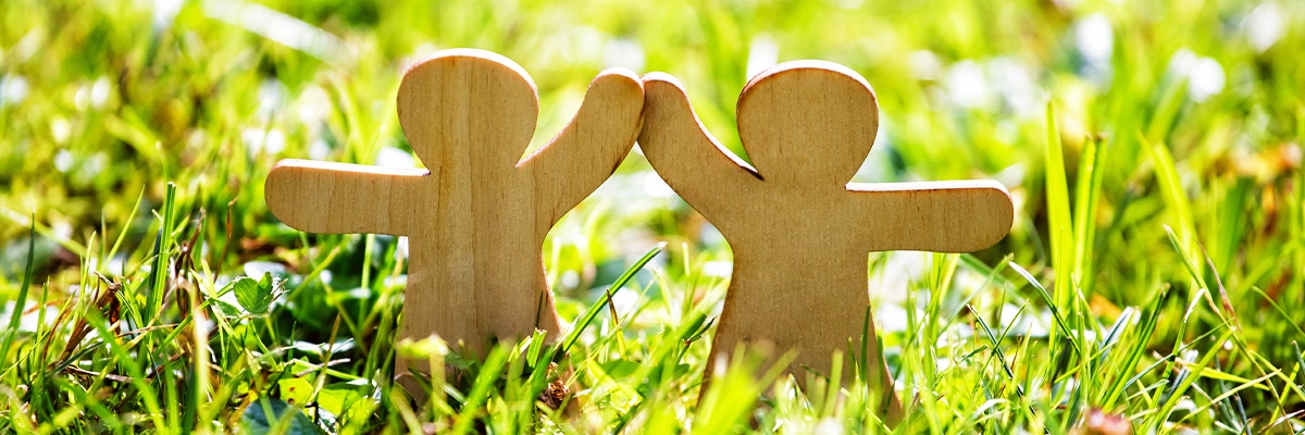Bild zeigt zwei Figuren aus Holz, welche auf einer sommerlichen Wiese stehen und sich an den Händen nehmen.