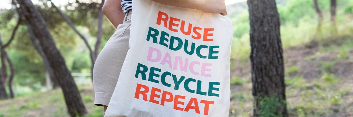 Die Abbildung zeigt eine im Wald spazierende Dame mit einer Stofftasche in der Hand. Darauf stehen die Worte: REUSE, REDUCE, DANCE, RECYCLE und REPEAT.