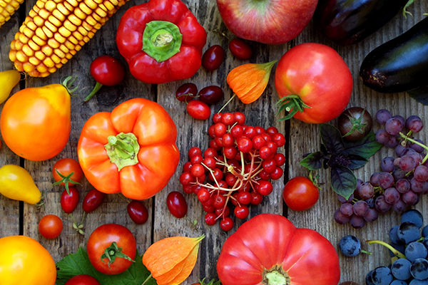 Abgebildet ist eine Vielzahl an Obst und Gemüse wie Paprika, Mais, Apfel und mehr auf einem Holztisch.