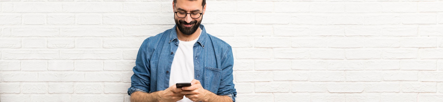 Ein Mann lehnt an einer weißen Ziegelwand und schaut auf sein Smartphone. 