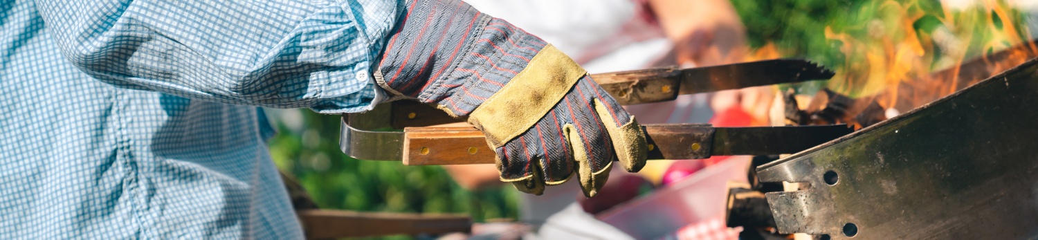 Ein Mann steht vor seinem Grill. Er trägt Schutzhandschuhe und hält eine Holzzange zum Wenden des Grillguts in seiner Hand.