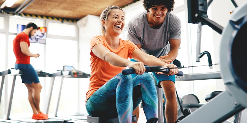 Eine lachende Frau im bekommt Fitnessstudio von einem schwarzhaarigen Mann eine Einführung am Rudergerät