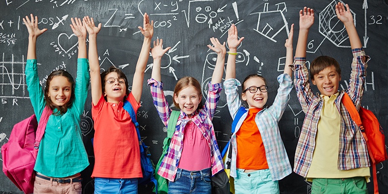 Fünf Kinder stehen vor einer Tafel mit erhobenen Händen, darauf sind Zeichnungen und Matheaufgaben.