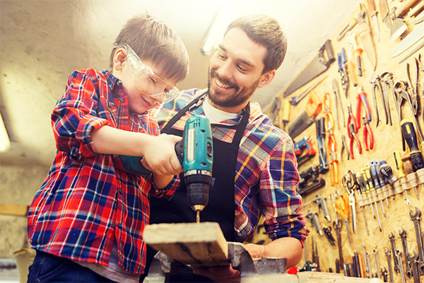 Vater und Sohn sind in der Werkstatt. Der Sohn trägt eine Brille und hält einen elektrischen Bohrer in der Hand.