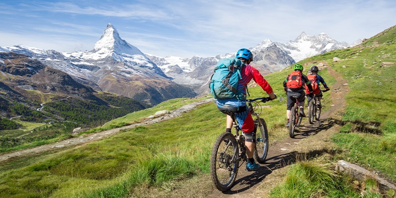 Drei Personen fahren Mountainbike auf einem Pfad in einer bergigen Gegend mit grüner Vegetation und schneebedeckten Gipfeln im Hintergrund.
