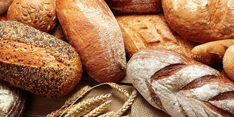 Frisches Brot in verschiedenen Formen und Texturen, mit Mehldekor und Weizenhalmen im Vordergrund.