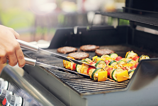 Auf dem Bild ist zu sehen, wie jemand einen Grillspieß auf einem Gasgrill wendet. Auf dem Gasgrill sind zudem Fleischpattys zu sehen.