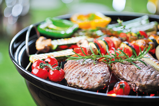 Auf dem Bild ist ein Kugelgrill mit diversen Grillgut ersichtlich. Auf dem Grill liegen Tomaten, Fleisch, Rosmarin, Paprika und vieles mehr.
