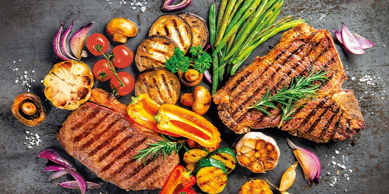 Gegrillte Steaks mit Gemüse wie Paprika, Pilzen, Tomaten und Spargel auf dunklem Untergrund.