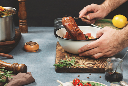 Auf dem Bild ist ersichtlich wie ein Fleisch mit einer Gabel aus der Marinade genommen wird. Rundherum sich Gewürze ersichtlich.