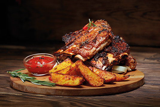 Auf dem Bild sind Spareribs mit Ketchup und Wedges auf einem Holzbrett ersichtlich. Garniert ist das Brett mit Rosmarin.
