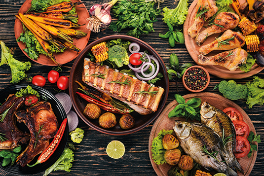 Auf dem Bild ist ein gedeckter Tisch mit fünf Tellern und verschiedenstem Grillgut wie zum Beispiel Fische, gegrillte Karotten oder Hühnerkeulen.