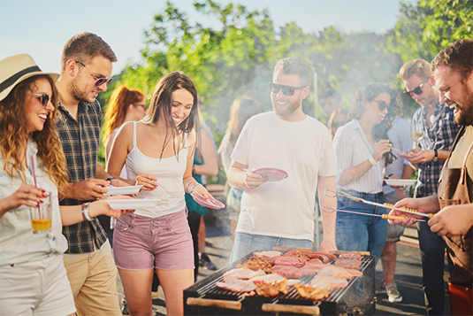Auf dem Bild sind fünf lächelnde, junge Erwachsene ersichtlich. Diese Personen stehen vor einem Grill mit verschiedensten Fleisch. Sie halte Teller in der Hand und eine Person hat auch ein Getränk. 
