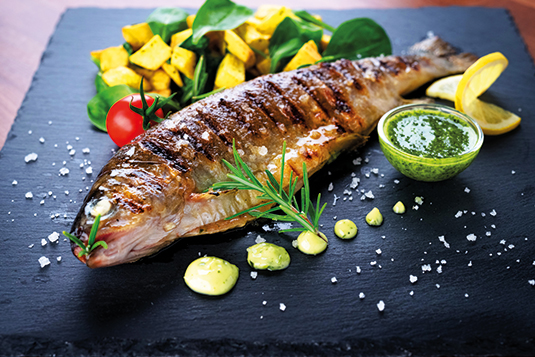 Auf dem Bild ist ein gegrillter Fisch auf einer Schieferplatte ersichtlich. Garniert wird mit Meersalz, gewürfelten Kartoffeln, Zitrone und einer Tomate.