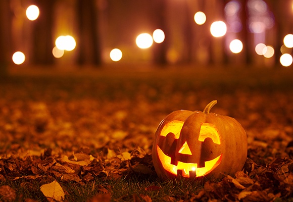 Geschnitzter Kürbis mit leuchtendem Gesicht zwischen Herbstlaub, unscharfe Lichter im Hintergrund.