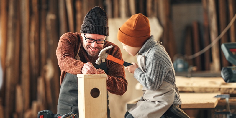 Zwei Personen arbeiten an einem Holzprojekt, eine hämmert einen Nagel in eine Box, Werkzeuge im Hintergrund.