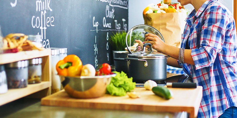 Person in kariertem Hemd kocht in einer Küche. Auf der Arbeitsfläche liegen Gemüse, ein Messer und eine Einkaufstasche.