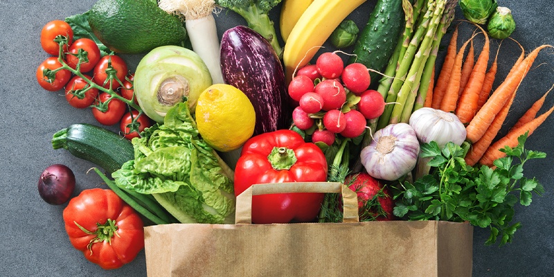 Frisches Gemüse und Obst in einer braunen Papiertüte, inklusive Tomaten, Blumenkohl, Bananen und Salat.
