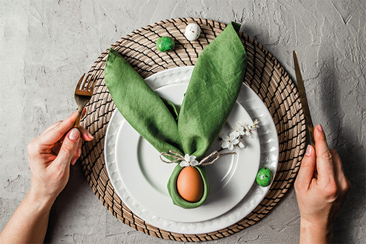 Um ein Ei wurde eine Serviette gelegt. Die Serviette sieht dadurch wie ein Osterhase aus. Die Serviette liegt auf einem Teller. Zudem sind Hände mit Besteck auf dem Bild ersichtlich.