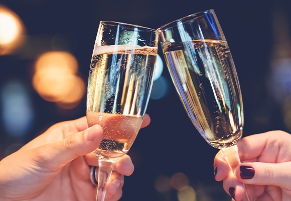 Zwei Hände stoßen mit Champagnergläsern an, unscharfes warmes Licht im Hintergrund.