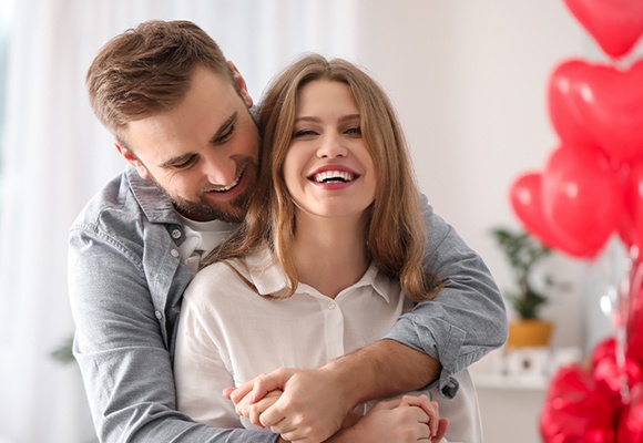 Zwei Personen stehen nah beieinander, eine Person umarmt die andere. Im Hintergrund sind rote Herzballons.