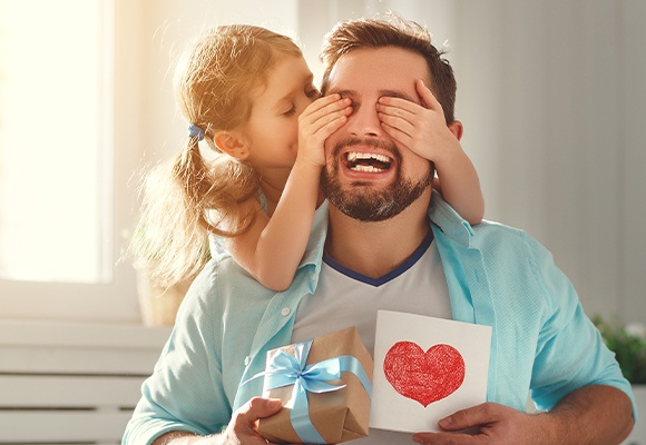 Person hält ein Geschenk mit braunem Papier und blauem Band, Kind umarmt sie von hinten.