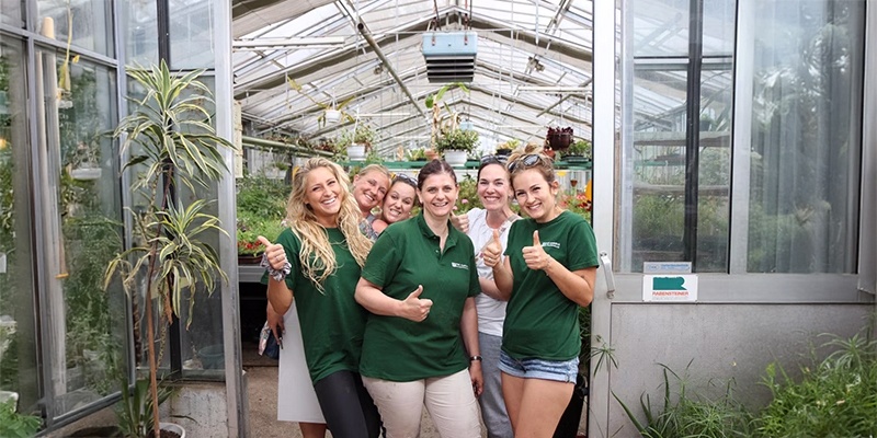 Man sieht eine Gruppe von Menschen, die den Daumen nach oben zeigen und in einem großen Glashaus stehen. 