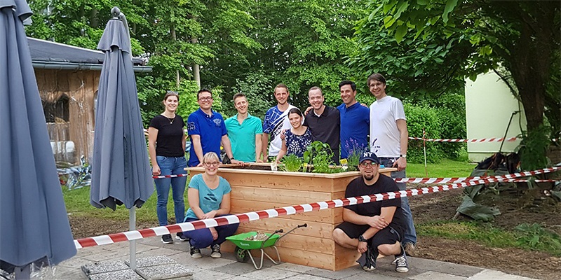 Man sieht eine Gruppe von Menschen, die in einem Garten gemeinsam neben einem Hochbeet stehen.