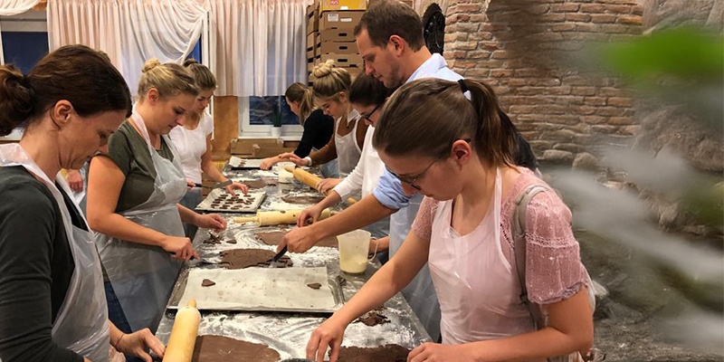 Man sieht acht Personen, die an einem langen Tisch Kekse backen. 