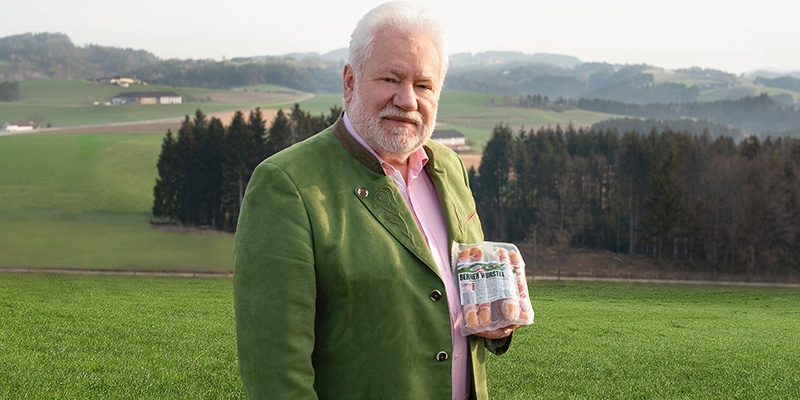 Herr mit grünem Sako steht auf einer Wiese mit einer Packung Würstel in der Hand.