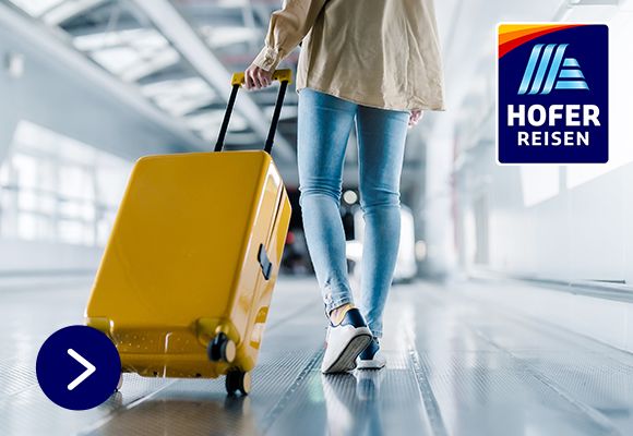 HOFER REISEN. Eine Person mit einer Jeans und Sneaker ist auf dem Flughafen hält einen gelben Reisekoffer in der Hand.