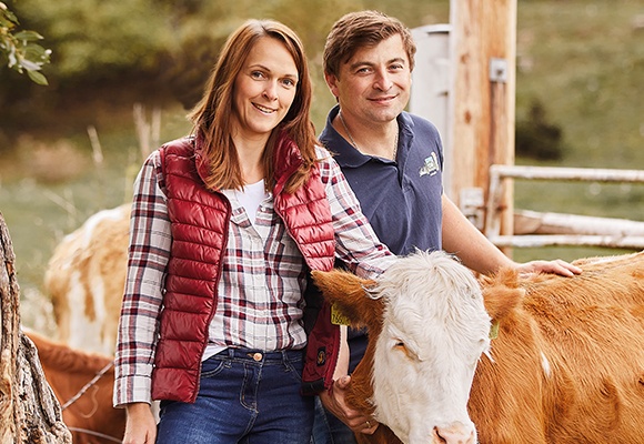 Eine Frau und ein Mann stehen in der Natur mit einer Kuh. Beide berühren die Kuh und lachen dabei.