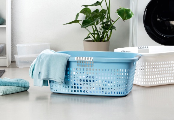 Am Boden stehen ein blauer und ein weißer Wäschekorb. Der blaue Wäschekorb ist mit Wäsche gefüllt. Im Hintergrund sieht man einen grünen Blumenstock, eine Waschmaschine und Aufbewahrungsboxen.