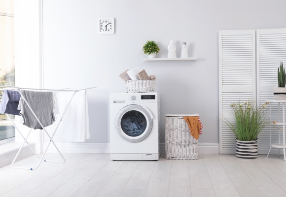 Wäscheständer mit aufgehänger Wäsche, daneben Waschmaschine, Wäschekorb, Blumentopf, kleiner Tisch und weißer Paravent, an der Mauer hängt ein Regal