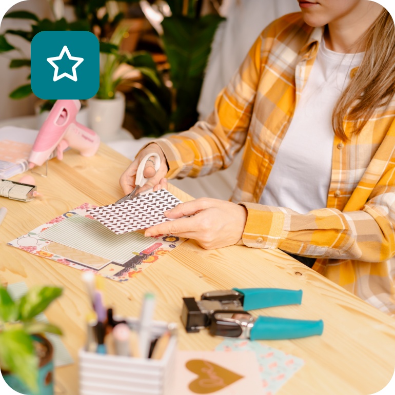 Frau in kariertem Hemd bastelt an einem Schreibtisch mit Papier, Schere und Bastelmaterialien. Kreative DIY-Handarbeitsszene.
