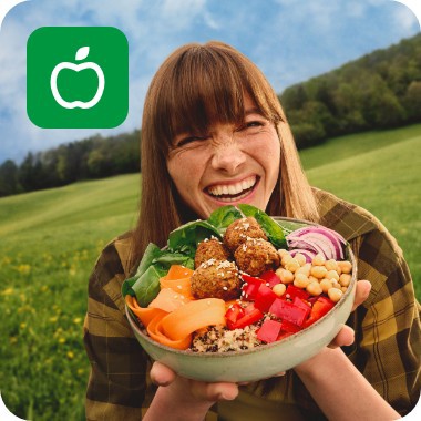 Junge Frau lacht fröhlich in der Natur und hält eine bunte Bowl mit frischem Gemüse, Falafel und Kichererbsen in den Händen.