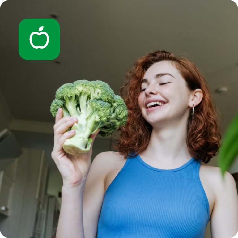 Lächelnde Frau in blauem Top hält einen Brokkoli in der Hand, während sie in ihrer Küche steht.