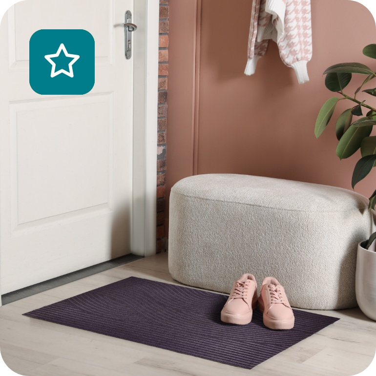 Eingangsbereich mit weißer Tür, ALDI Fußmatte in Anthrazit, rosa Schuhen, weißem Hocker und grüner Zimmerpflanze.