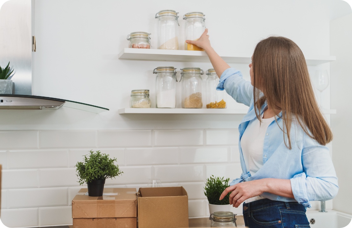 Frau in einer hellen Küche immt ein Glas mit Lebensmitteln von einem weißen Wandregal.