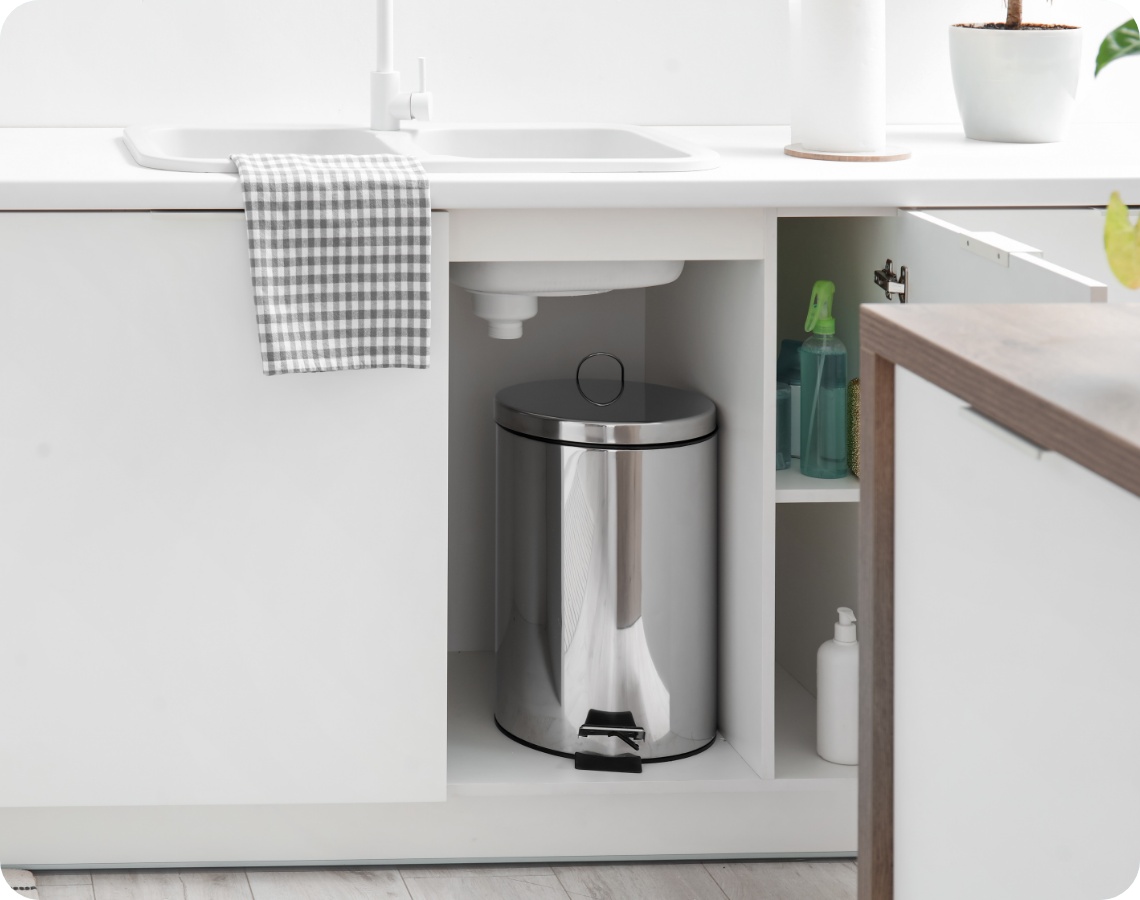 Geöffneter Küchenschrank unter der Spüle mit Edelstahl-Mülleimer und Putzmitteln in einer weißen, modernen Küche.