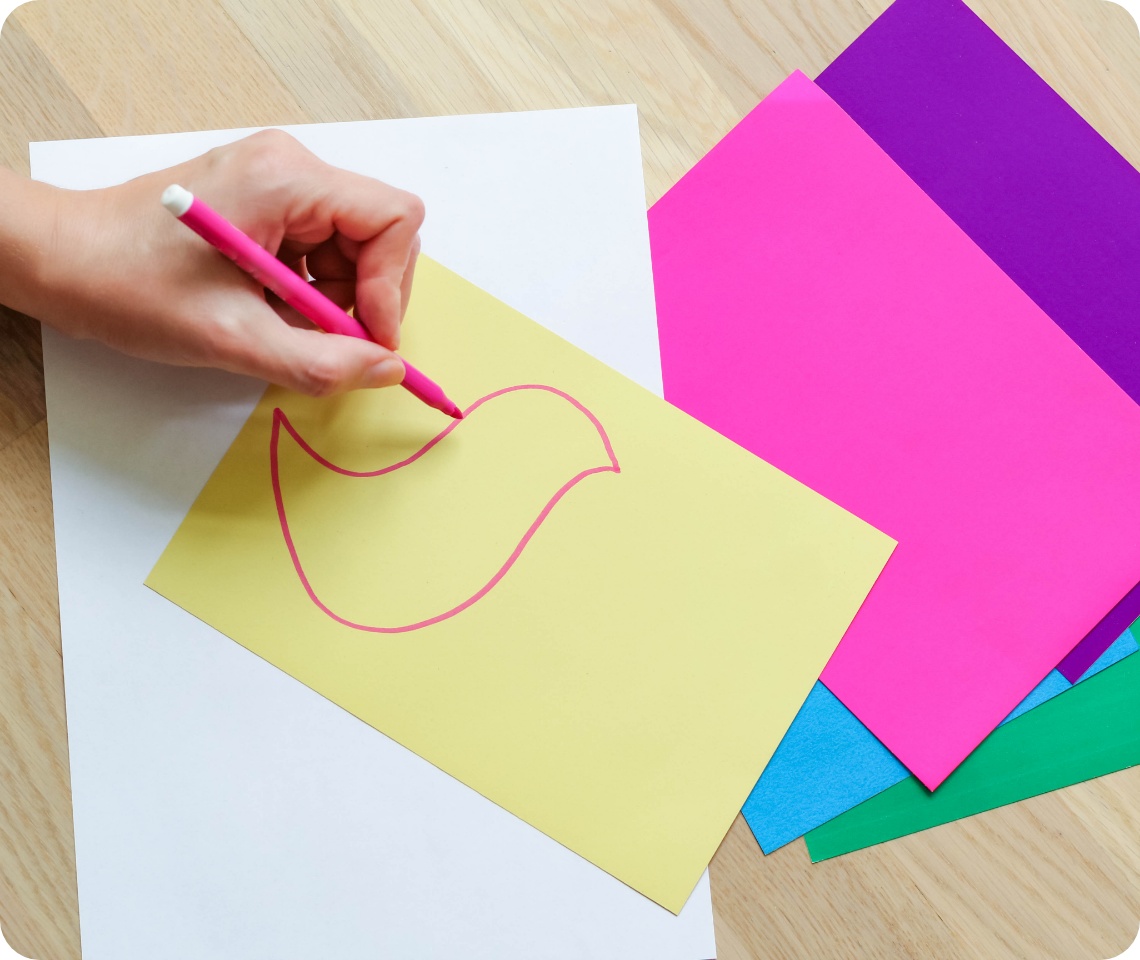 Hand zeichnet mit pinkem Stift eine Vogelkontur auf gelbes Bastelpapier. Bunte Papierbögen liegen auf einem Holztisch.