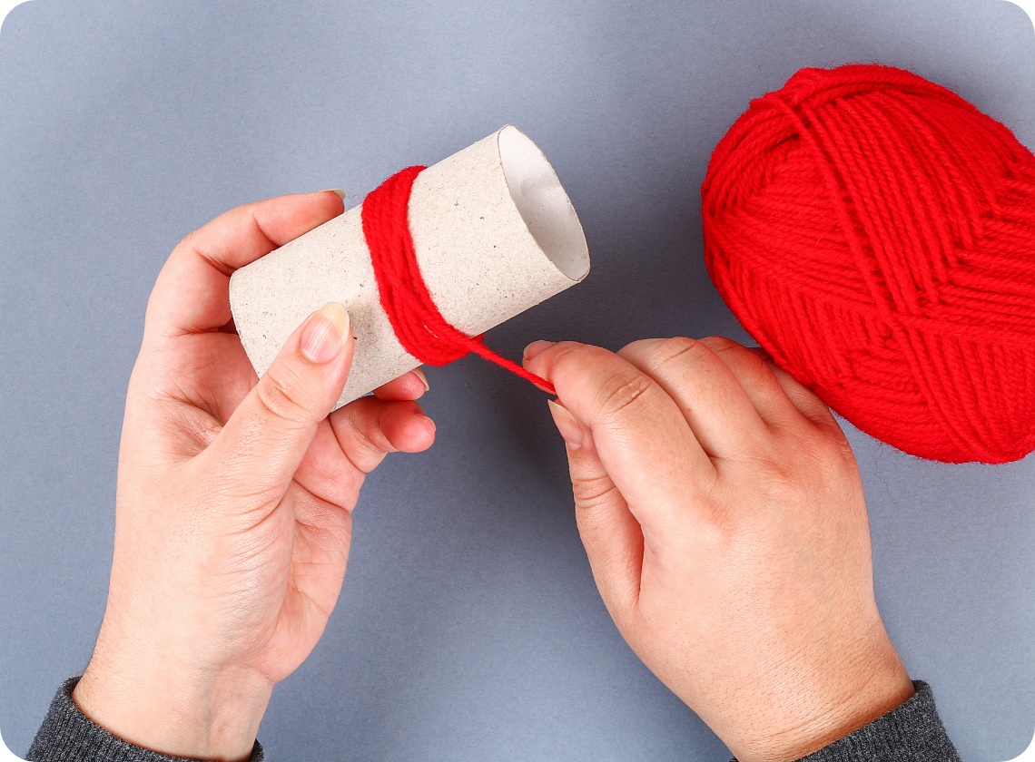 Hände wickeln rote Wolle um eine Papprolle, daneben ein Wollknäuel auf grauem Hintergrund. DIY-Bastelprojekt.