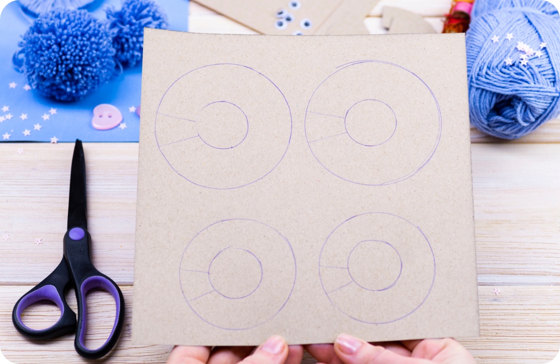Pappscheibe mit Kreisen, Schere, blaue Wolle und fertige Pompons auf Holztisch.